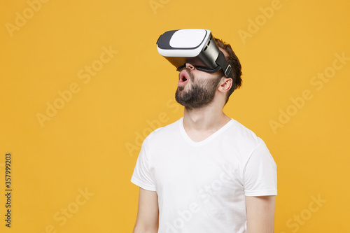Shocked young bearded man guy in white casual t-shirt posing isolated on yellow wall background studio portrait. People sincere emotions lifestyle concept. Mock up copy space. Watching in headset.