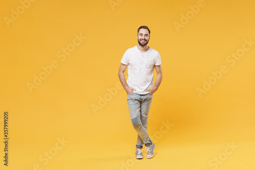Smiling young bearded man guy in white casual t-shirt posing isolated on yellow background studio portrait. People sincere emotions lifestyle concept. Mock up copy space. Holding hands in pockets.