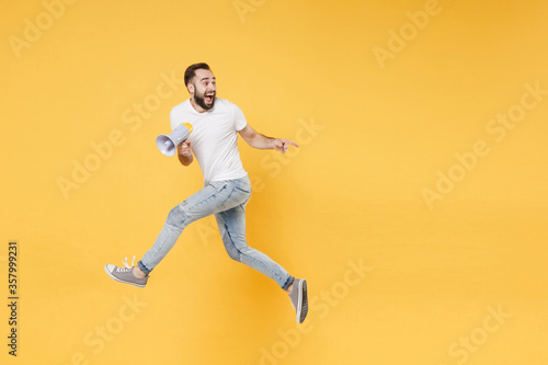 Excited young bearded man guy in white casual t-shirt posing isolated on yellow background studio. People lifestyle concept. Mock up copy space. Jumping  hold megaphone  pointing index finger aside.
