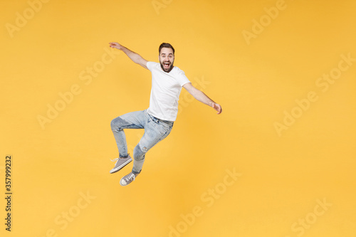 Excited young bearded man guy in white casual t-shirt posing isolated on yellow wall background studio portrait. People sincere emotions lifestyle concept. Mock up copy space. Jumping spreading hands. © ViDi Studio
