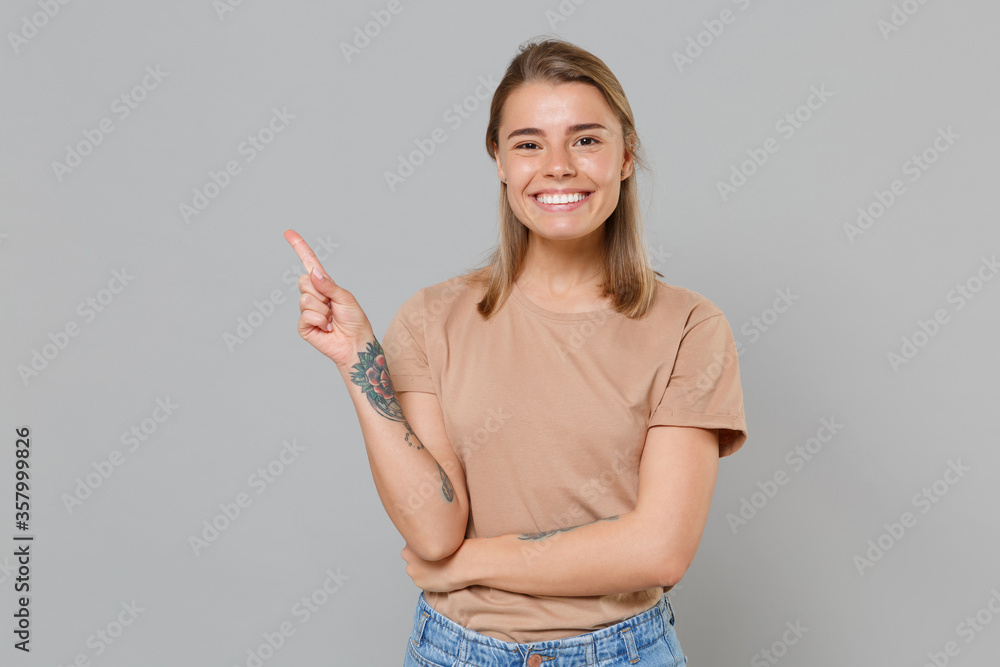 Smiling young blonde woman girl in casual beige t-shirt posing isolated on gray wall background studio portrait. People emotions lifestyle concept. Mock up copy space. Pointing index finger aside up. Smiling young blonde woman girl in casual beige t-shirt posing isolated on gray wall background studio portrait. People emotions lifestyle concept. Mock up copy space. Pointing index finger aside up.