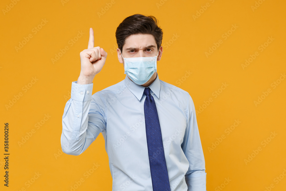Displeased young business man in classic blue shirt tie sterile face mask isolated on yellow background. Epidemic pandemic coronavirus 2019-ncov sars covid-19 flu virus concept. Point index finger up. Displeased young business man in classic blue shirt tie sterile face mask isolated on yellow background. Epidemic pandemic coronavirus 2019-ncov sars covid-19 flu virus concept. Point index finger up.