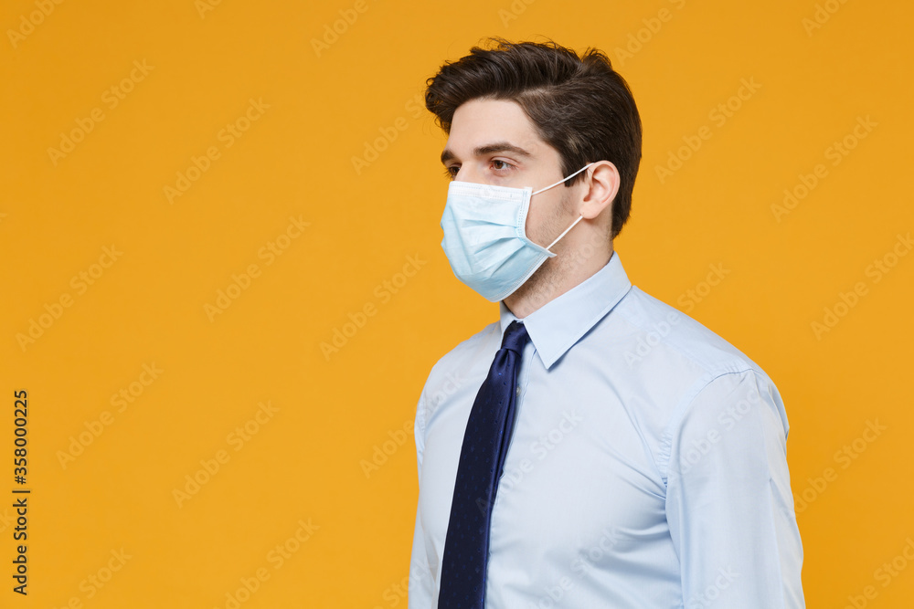 Young business man in classic blue shirt tie sterile face mask isolated on yellow background. Epidemic pandemic rapidly spreading coronavirus 2019-ncov sars covid-19 flu virus concept. Looking aside.