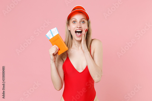 Shocked blonde woman in red one-piece swimsuit cap isolated on pink background. People summer vacation rest lifestyle concept. Air flight journey. Hold passport ticket boarding pass put hand on cheek. © ViDi Studio
