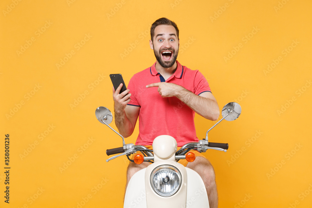 Surprised young bearded guy in casual summer clothes driving moped isolated on yellow background. Driving motorbike transportation concept. Mock up copy space. Pointing index finger on mobile phone.