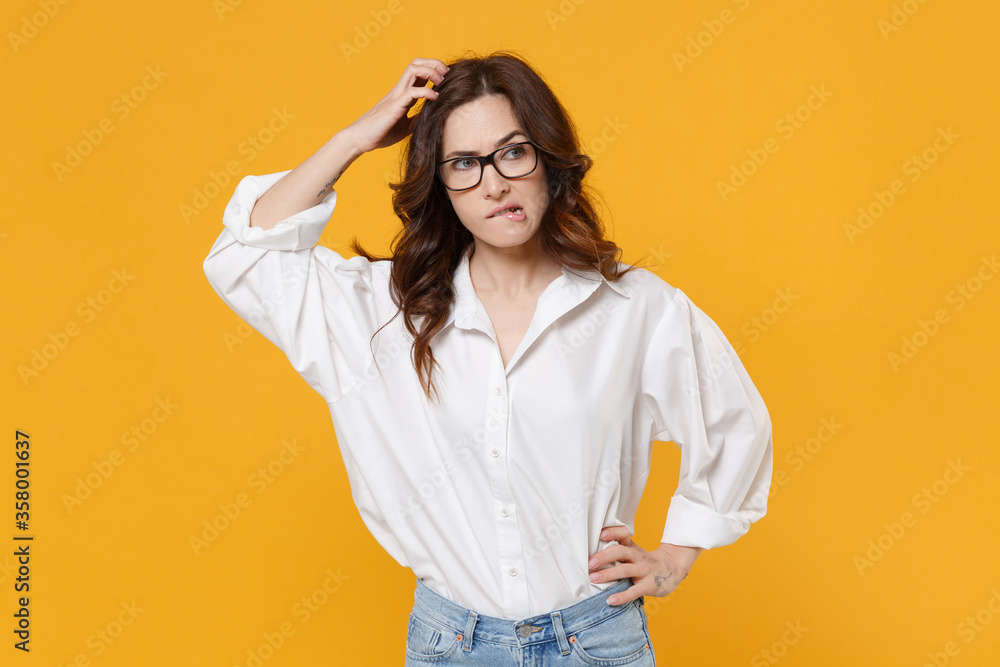 Preoccupied young brunette business woman in white shirt glasses isolated on yellow background studio. Achievement career wealth business concept. Mock up copy space. Put hand on head, biting lips. Preoccupied young brunette business woman in white shirt glasses isolated on yellow background studio. Achievement career wealth business concept. Mock up copy space. Put hand on head, biting lips.