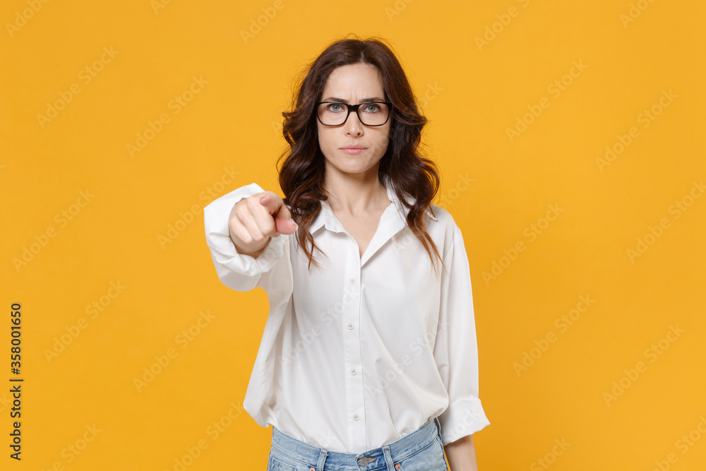 Strict young brunette business woman in white shirt glasses isolated on yellow wall background studio. Achievement career wealth business concept. Mock up copy space. Pointing index finger on camera.