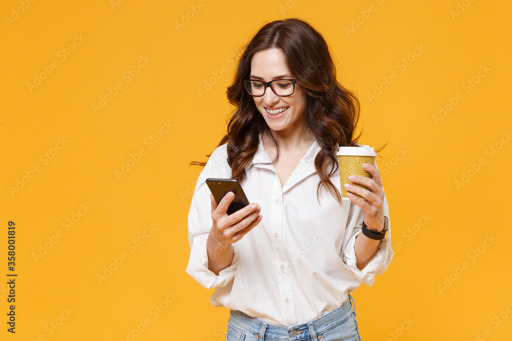 Smiling young business woman in white shirt glasses isolated on yellow background. Achievement career wealth business concept. Mock up copy space. Using mobile phone, hold paper cup of coffee or tea. Smiling young business woman in white shirt glasses isolated on yellow background. Achievement career wealth business concept. Mock up copy space. Using mobile phone, hold paper cup of coffee or tea.