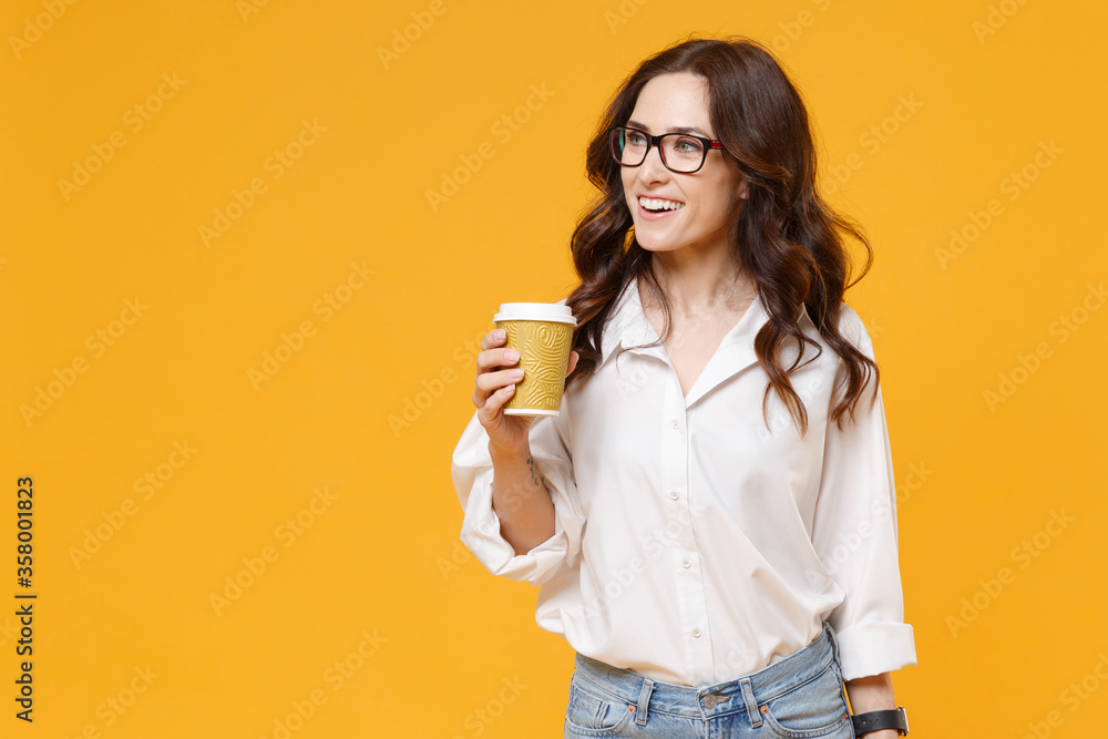 Smiling young business woman in white shirt glasses isolated on yellow wall background. Achievement career wealth business concept. Mock up copy space. Hold paper cup of coffee or tea, looking aside. - obrazy, fototapety, plakaty 