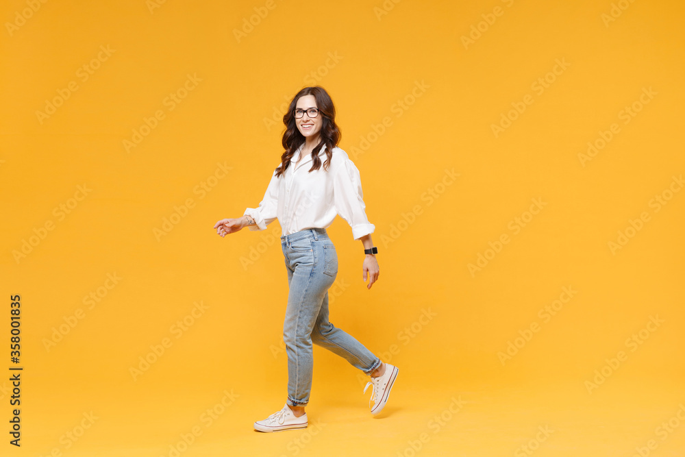 Side view of smiling young brunette business woman in white shirt glasses isolated on yellow background. Achievement career wealth business concept. Mock up copy space. Walking going, looking camera. Side view of smiling young brunette business woman in white shirt glasses isolated on yellow background. Achievement career wealth business concept. Mock up copy space. Walking going, looking camera.
