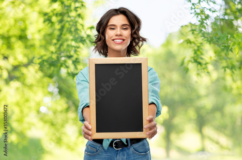 people concept - portrait of happy smiling young woman in turquoise shirt showing black chalkboard over green natural background