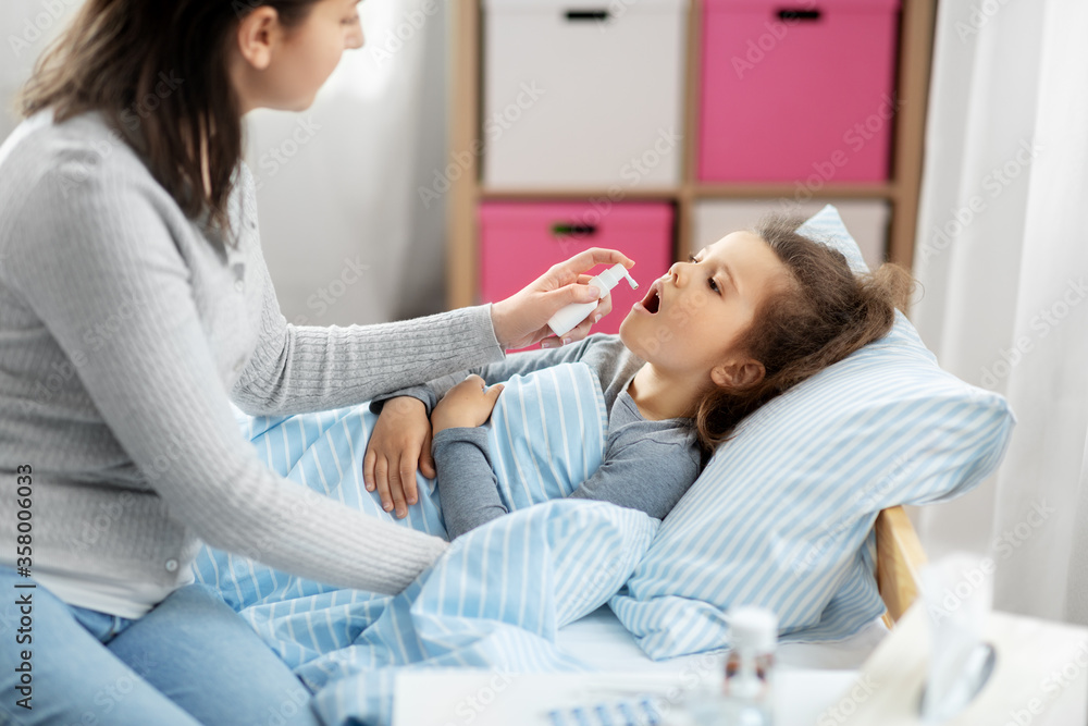 Fototapeta premium family, health and medicine concept - mother with oral spray treats little sick daughter lying in bed at home