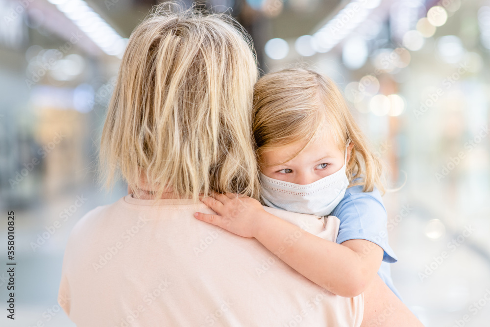 Sad baby girl wearing medical protective mask hugs mother in a public crowded place - in a shopping mall or airport Sad baby girl wearing medical protective mask hugs mother in a public crowded place - in a shopping mall or airport
