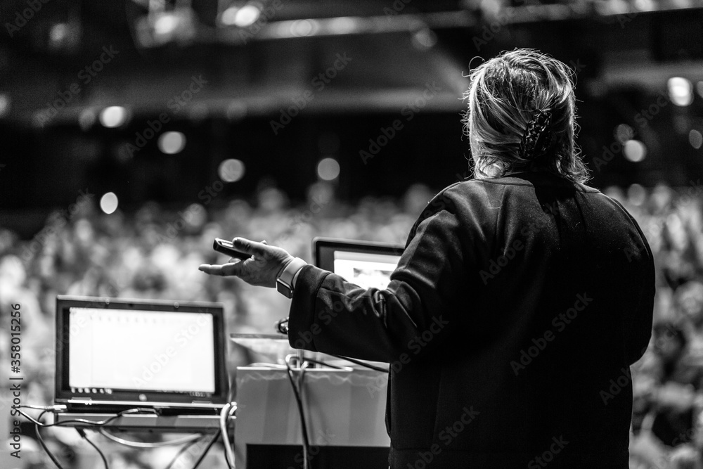 Female speaker giving a talk on corporate business conference. Unrecognizable people in audience at conference hall. Business and Entrepreneurship event. Black and white image. Female speaker giving a talk on corporate business conference. Unrecognizable people in audience at conference hall. Business and Entrepreneurship event. Black and white image.