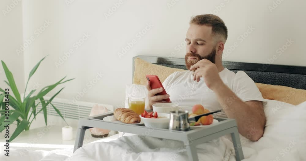 Good looking bearded man using his mobile phone and eating while sitting on bed. Handsome man in 30s having breakfast while sitting with tray full of food and looking at phone screen. Good looking bearded man using his mobile phone and eating while sitting on bed. Handsome man in 30s having breakfast while sitting with tray full of food and looking at phone screen.
