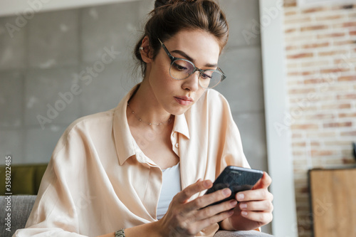 Image of thinking woman using mobile phone while sitting on couch