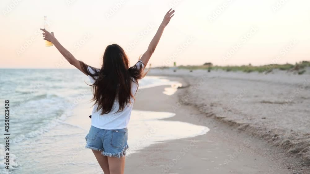 Pretty woman walking along seaside on sandy beach having fun with beer bottle. Attractive fitted girl raises hands, enjoys fredom and summer vacation near ocean shore, drinking beverage. Slow motion. Pretty woman walking along seaside on sandy beach having fun with beer bottle. Attractive fitted girl raises hands, enjoys fredom and summer vacation near ocean shore, drinking beverage. Slow motion.