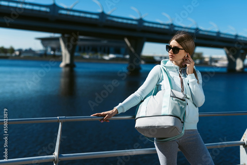 A young woman with a sports bag, in a tracksuit, walking along the promenade, listening music and preparing for street sports.