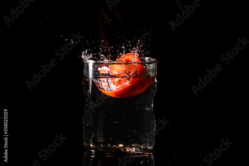 A glass of water in a dark background and water being splashed with a piece of cut tomato dropped inside the glass