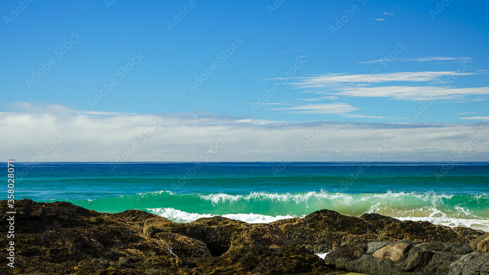 Looking across a blue-green ocean, with waves breaking onto rocks in the foreground, and clouds on the horizon, beneath a blue sky: at Currumbin Beach, Gold Coast, Queensland, Australia.