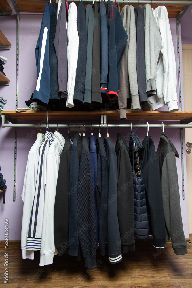 men's shirts, jackets and sweaters of different calm dark colors hang on the bar in the wardrobe. wooden shelves. lilac wall