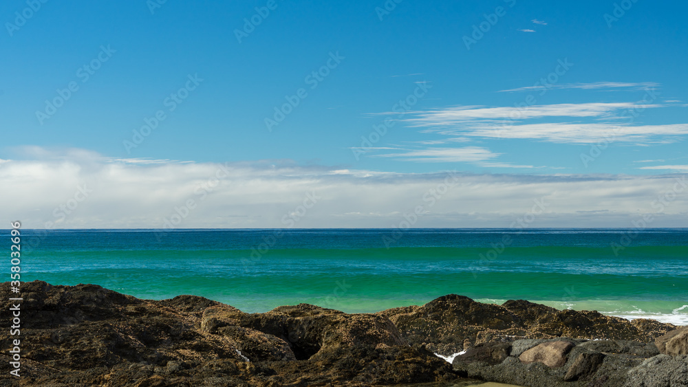Seascape with blue-green waves rising a beyond rocky foreground, and clouds on the horizon, beneath a blue sky. At Currumbin, Gold Coast, Queensland, Australia.