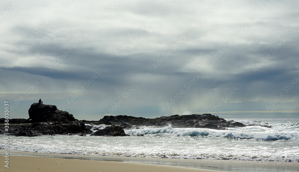 Storm over the sea watched by man and seagulls sitting on a rock, with waves breaking on the sand in the foreground. Currumbin Beach, Gold Coast, Queensland, Australia.