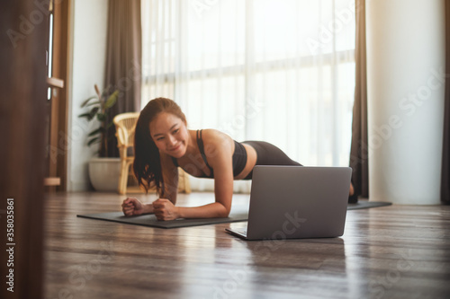 A beautiful young asian woman doing plank on training mat while watching online workout tutorials on laptop at home