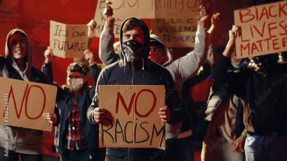 Φωτογραφία Caucasian young man in hood standing in crowd of people at street protest with poster No racism at night
