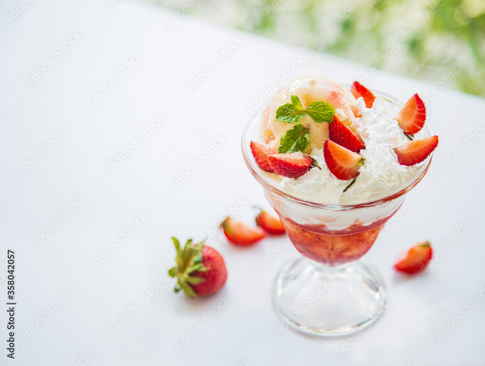 Close up side views a cup of delicious strawberries sundae with mint leaves on a white table next to the glass window through nature background in the afternoon.