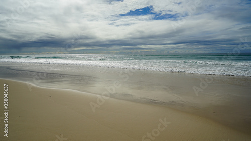 Expansive view from sandy beach across breaking waves to low clouds and rain on the horizon, at Currumbin Beach, Gold Coast, Queensland, Australia.