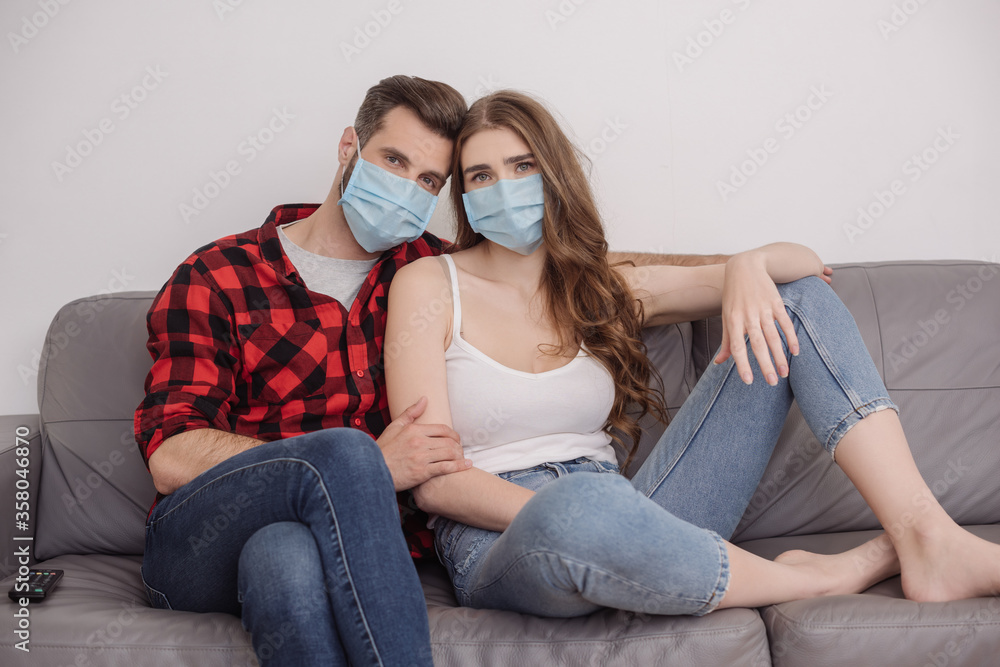depressed young couple in medical masks sitting on sofa and looking at camera
