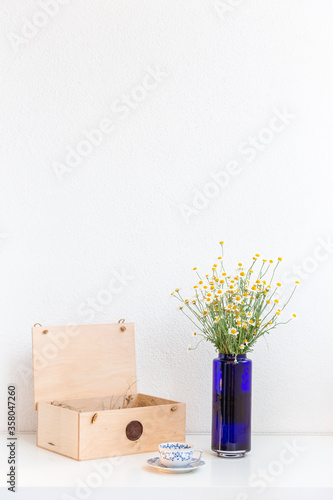 Bouquet of daisies in a blue vase with vintage tea cup and wooden box on white background. Still-life. With copy space. Cup of aromatic tea with a camomile flowers. Place for text. 