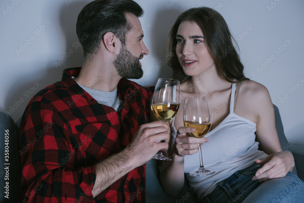 happy young couple sitting on sofa, looking at each other while holding glasses of white wine happy young couple sitting on sofa, looking at each other while holding glasses of white wine