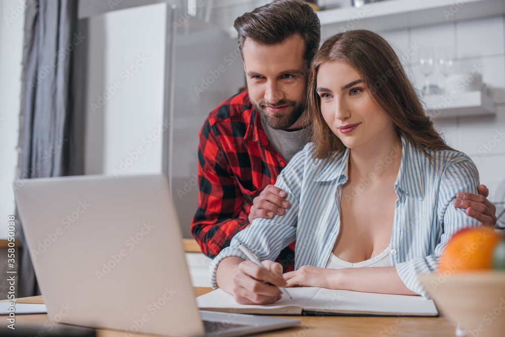 selective focus of smiling man touching shoulders of pretty girlfriend working at laptop selective focus of smiling man touching shoulders of pretty girlfriend working at laptop