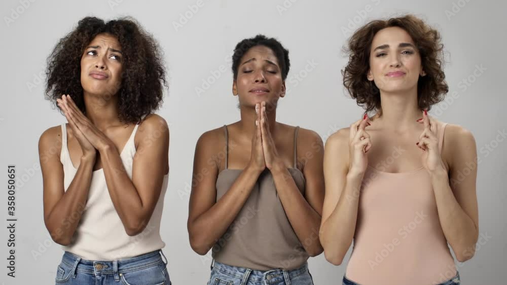 Three beautiful multi-ethnic women are making a wish while standing with crossed fingers isolated over white background