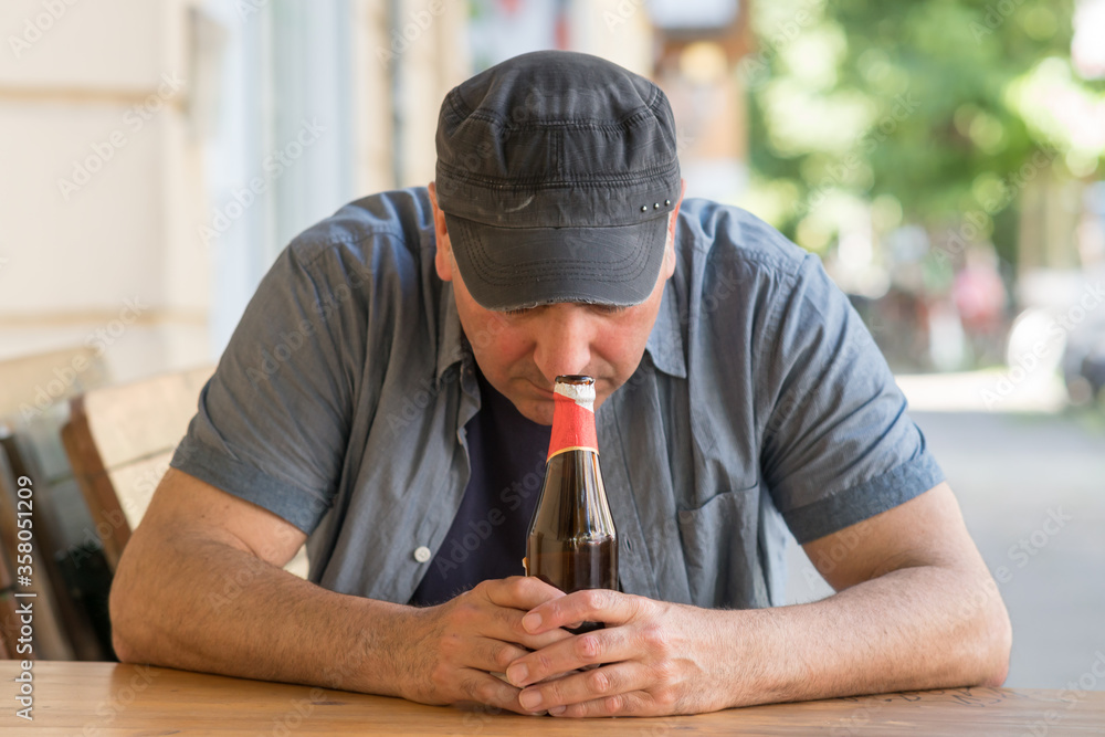 Pensive and sad man with bowed head sitting holding a bottle of beer outside a bar. Selective focus