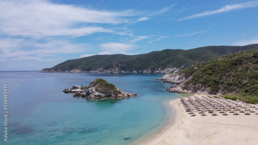 Mediterranean terrain Greek landscape sandy beach drone shot. Aerial view of Sithonia Chalkidiki peninsula with green plantation by crystal clear waters, sea beds with umbrellas & small rocky island.