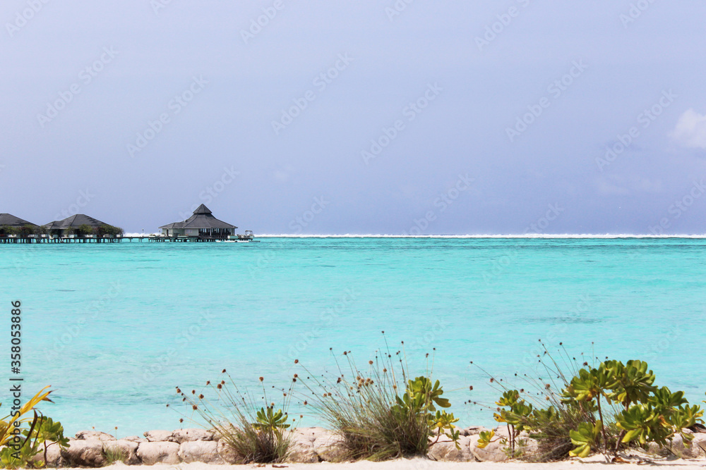 Obraz premium Azure waters of Indian waters on foreground, wooden open air restaurant on the background, summer vacation on Maldives islands. Luxury resort, holiday and travel concept