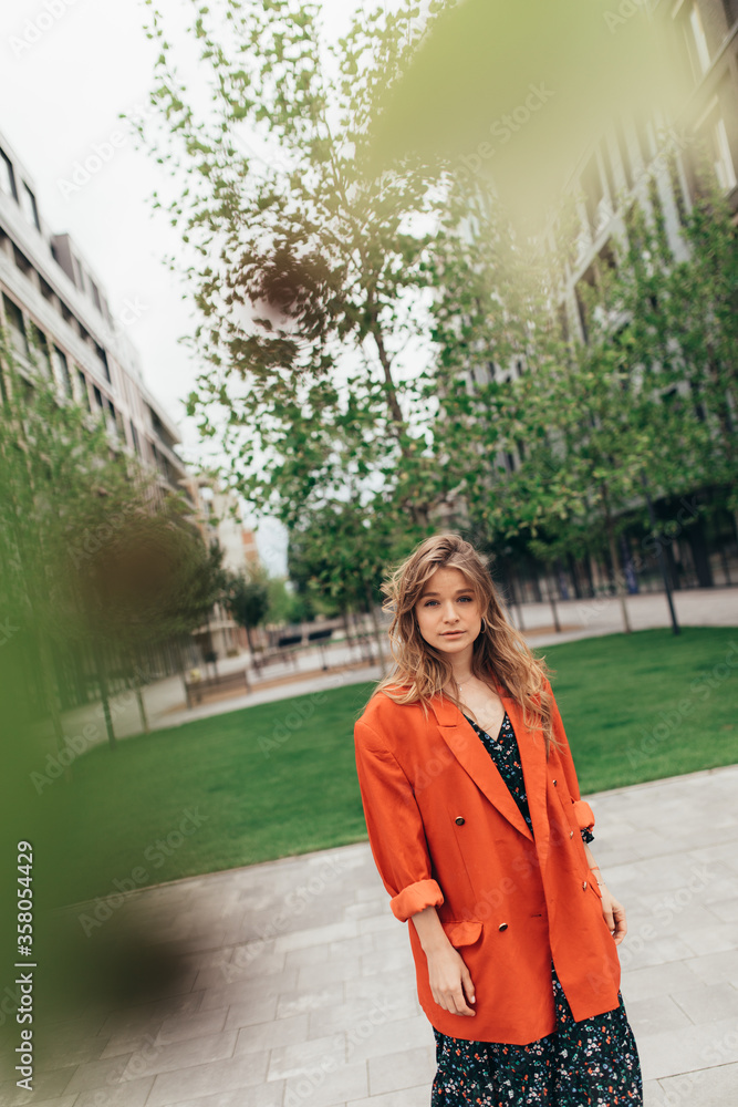 Picture is made at angle. Young stylish trendy woman posing on camera hidden behind tree leaves. Girl wears dark dress and orange jacket. Fashionable picture on the street.