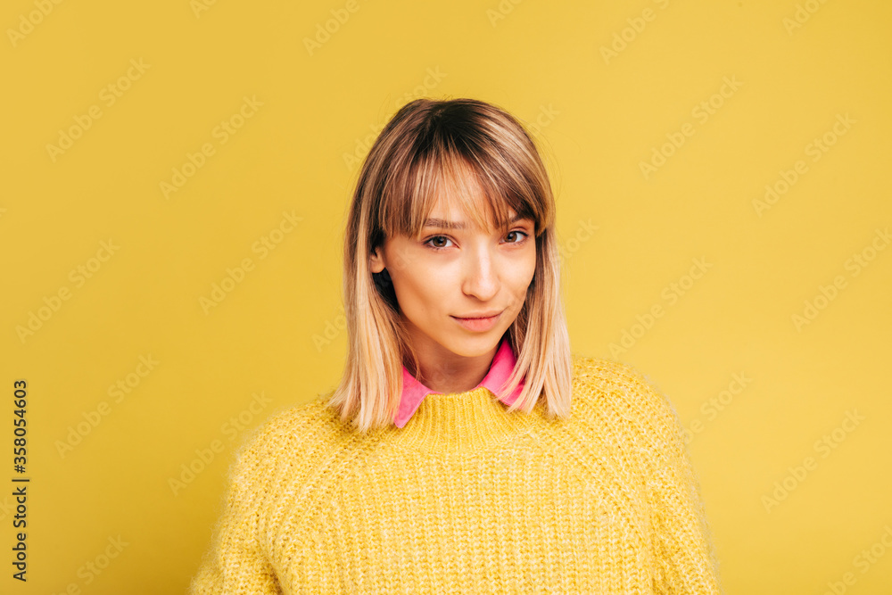 Confident beautiful young woman looks straight and pose on camera. Blonde girl wear yellow sweater with pink collar. Stand alone in studio. Isolated over yellow background. Confident beautiful young woman looks straight and pose on camera. Blonde girl wear yellow sweater with pink collar. Stand alone in studio. Isolated over yellow background.