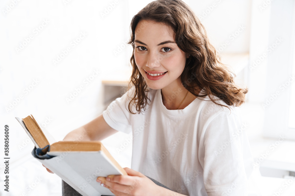 Photo of pleased woman smiling and reading book while sitting on bed Photo of pleased woman smiling and reading book while sitting on bed