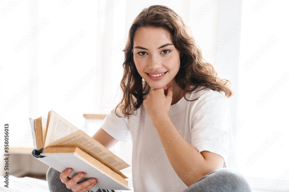 Photo of pleased woman smiling and reading book while sitting on bed Photo of pleased woman smiling and reading book while sitting on bed