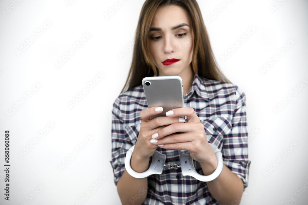 brunette young beautiful concentrated woman holds smartphone in her hands, texts while having handcuffs on , virtual reality concept, social media addiction, nomophobia, isolated white background brunette young beautiful concentrated woman holds smartphone in her hands, texts while having handcuffs on , virtual reality concept, social media addiction, nomophobia, isolated white background