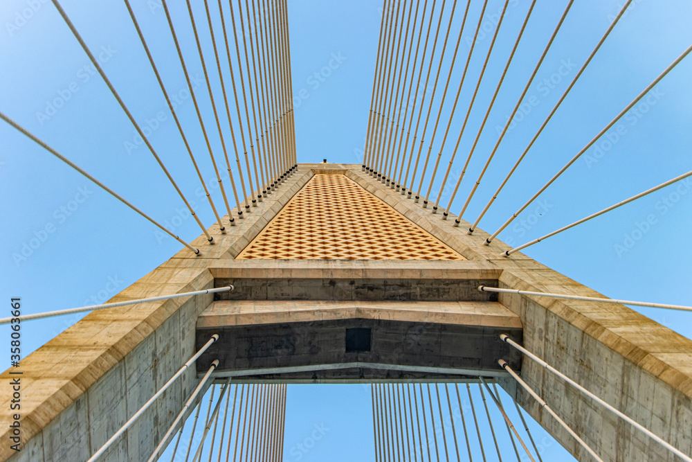 Fototapeta premium Concrete bridge pylon with parallel lines of steel cable. Passage under the supporting tower of the cable bridge. Construction Cable-stayed bridge against blue sky.