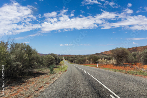 Road at the Red centre of Australia. Empty road, no cars, no signs. Green vegetation and bush on the sides. Orange and red soil. MacDonnell ranges, Northern Territory NT, Australia, Oceania