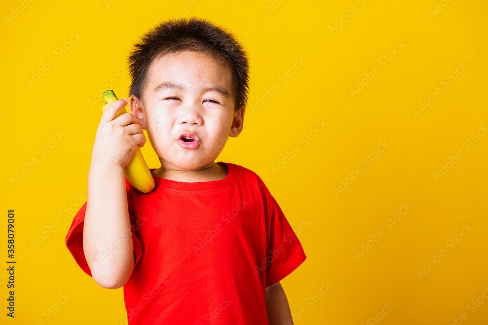 Happy portrait Asian child or kid cute little boy attractive smile wearing red t-shirt playing holds banana fruit pretending to be like a telephone, studio shot isolated on yellow background Happy portrait Asian child or kid cute little boy attractive smile wearing red t-shirt playing holds banana fruit pretending to be like a telephone, studio shot isolated on yellow background
