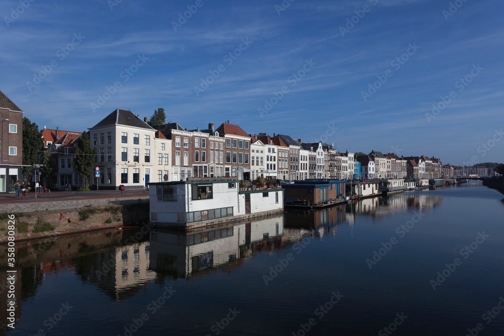 Floating home boats in front of historic mansions reflected in a canal in Middelburg, The Netherlands against a blue sky with streak clouds