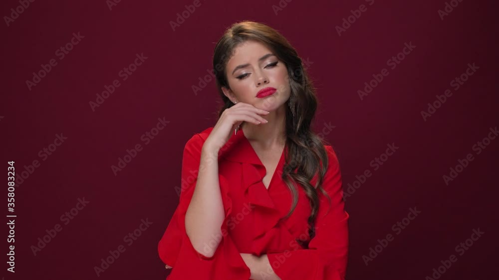 An upset woman wearing red dress is looking to the camera isolated over burgundy background An upset woman wearing red dress is looking to the camera isolated over burgundy background