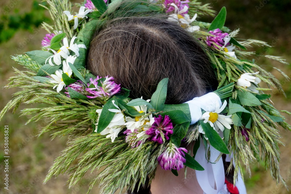 Naklejka premium Beautiful woman in wreath of midsummer flowers on heads in meadow on sunny summer day. rear view 